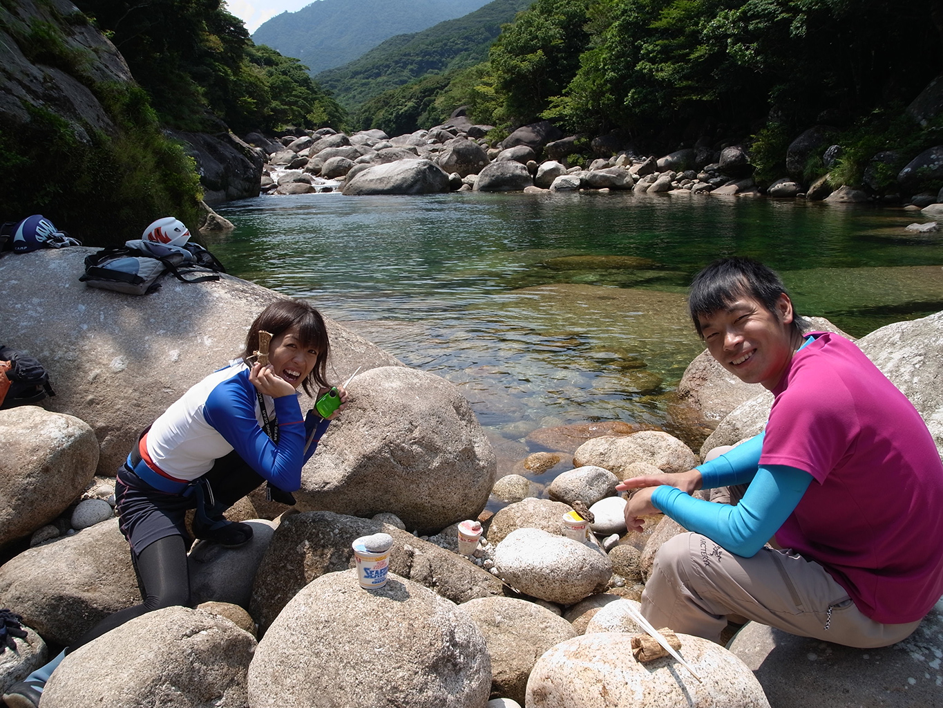 川岸でお昼を食べている写真。軽めのお弁当の後は、〆のカップラーメンを食べる予定で、カップヌードルの上に石を置いて完成を待っている写真