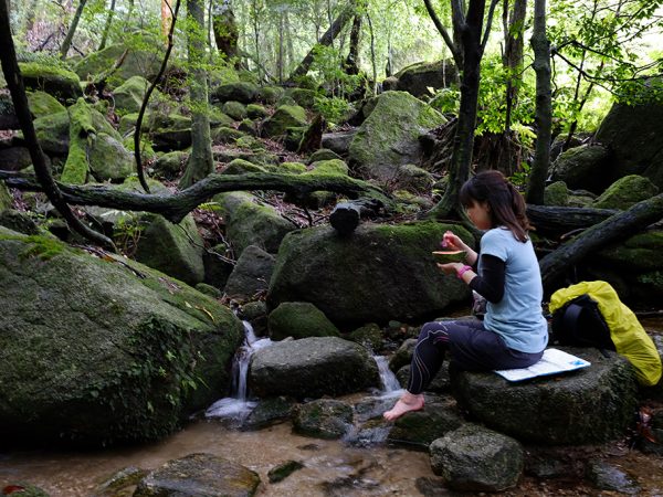 美しい苔と沢の景色を眺めながらスイカを頬張る参加者Kさんの写真