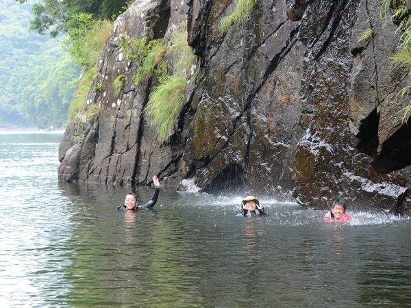 岩壁から落ちてくる石清水を水に浮かびながら体で受け止めて遊んでいる3人の写真