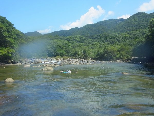 奥には山の緑と青空が映え、そこを流れる静かな流れに仰向けになって流れてくる二人の写真