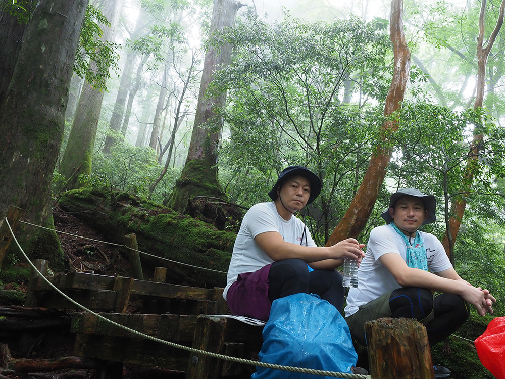 雨がポツポツとする中深い森の中で休憩する２人の写真